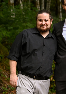 A smiling man in a black shirt stands in the woods.
