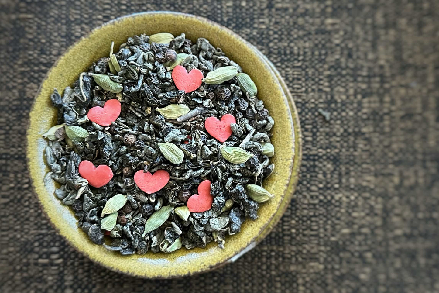 Green teacup full of rolled green and oolong tea with cardamom pods, black peppercorn and red heart sprinkles