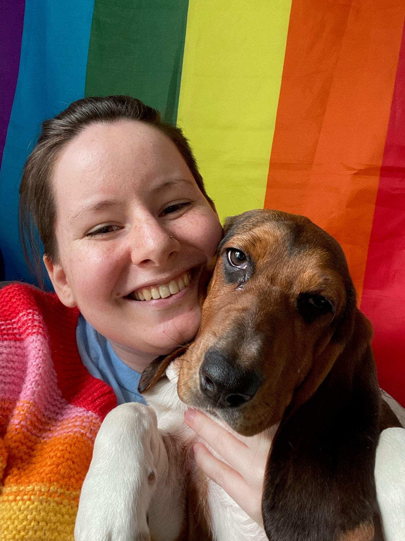 A smiling human hugs a basset hound. A rainbow flag hangs in the background.