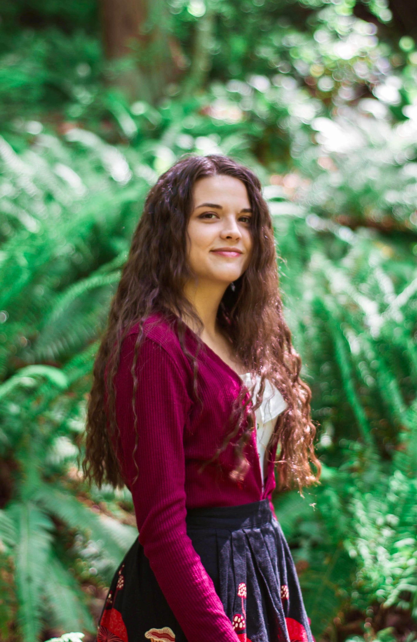 A smiling human in a forest with long brown wavy hair and a red cardigan.