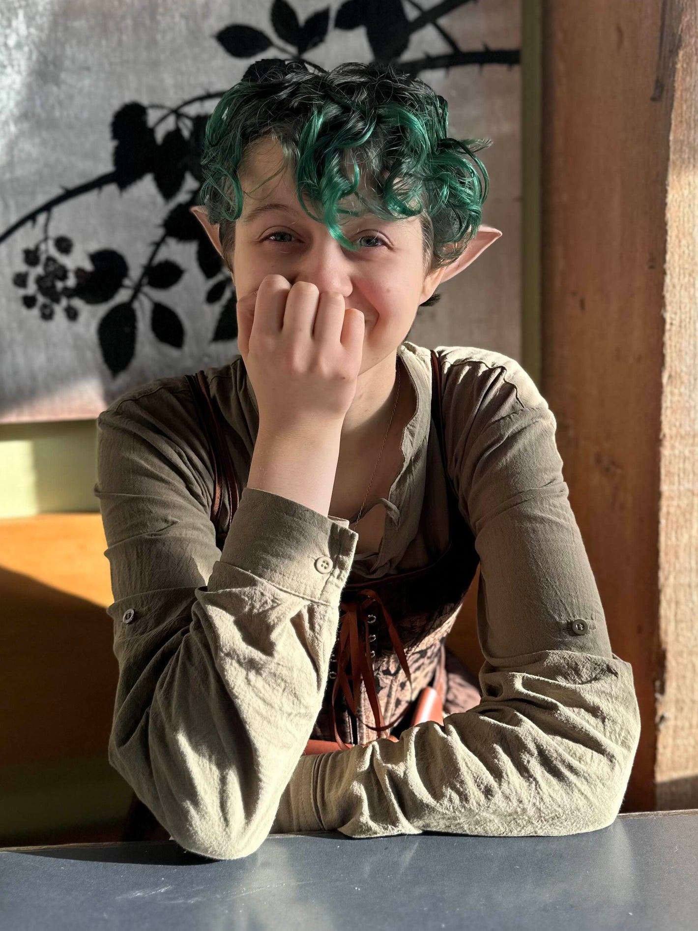 Person with green hair sitting at a table in a room with a painting on the wall.