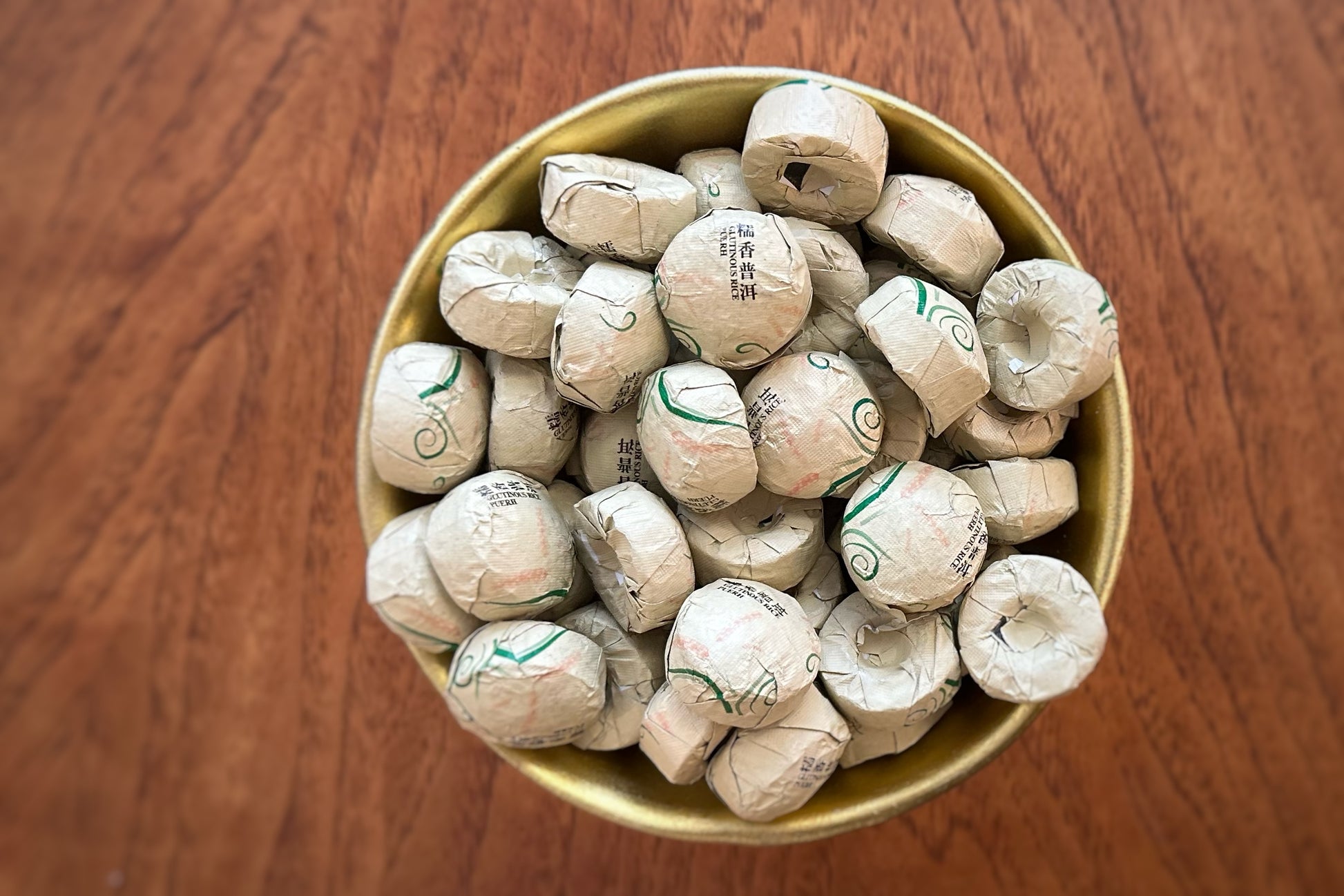 Gold bowl filled with small foil-wrapped tea cakes on a wooden surface