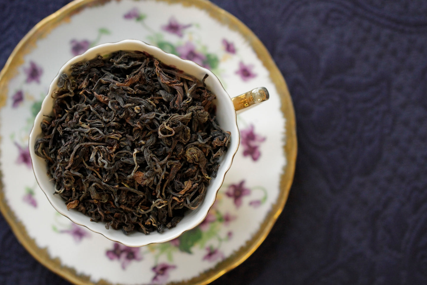 A cup of dry oolong tea leaves placed on a saucer with floral patterns, on a dark textured background.