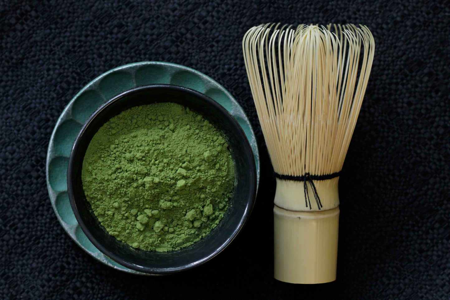A matcha whisk sits beside a cup of loose green tea powder