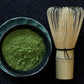 A teacup full of matcha powder beside a bamboo whisk